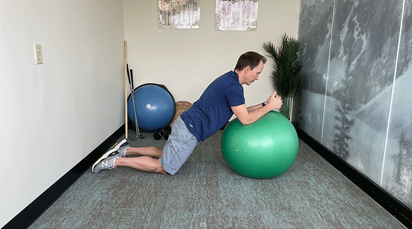A man with brown hair, a blue sports shirt, and light grey shorts performs an ab roll out with his knees on the floor and his upper body lifted onto a green stability ball.