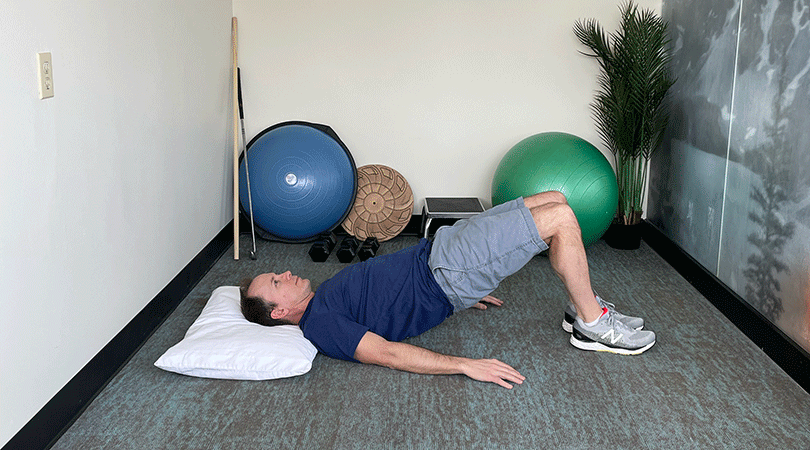A man with brown hair, a blue sports t-shirt, and grey shorts performs a bridge exercise with his head and both feet planted on the ground, raising his hips toward the ceiling.