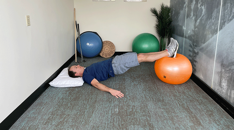A man with brown hair performing a hamstring curl by laying on his back with his legs raised and lifted onto and orange exercise ball.