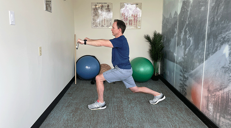 A man with brown hair, a blue sports t-shirt, and grey shorts is standing in a lunch with the forward facing leg out infront of him and bent at the knee. He holds a golf club horizontally between both hands and twists his torso from side to side.