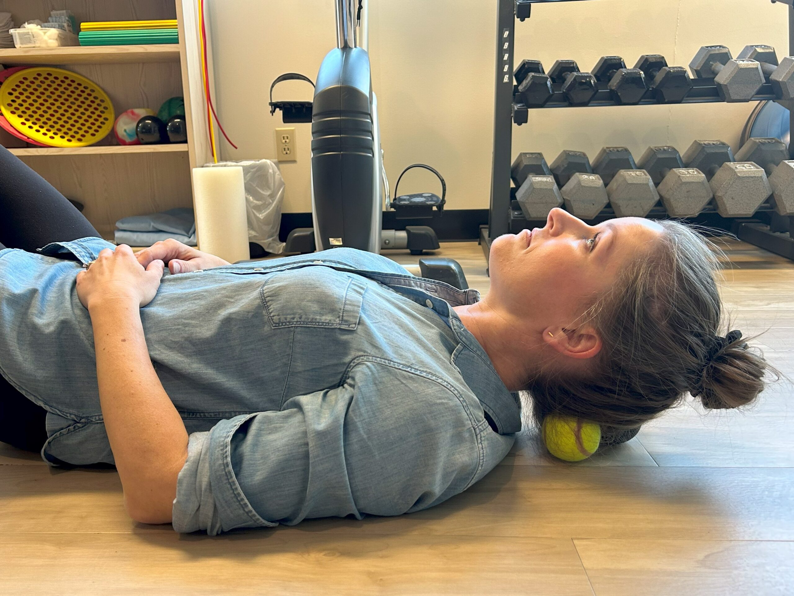 A lady is laying on the ground with a tennis ball behind her upper neck. She uses the ball to roll out the tension on her neck.