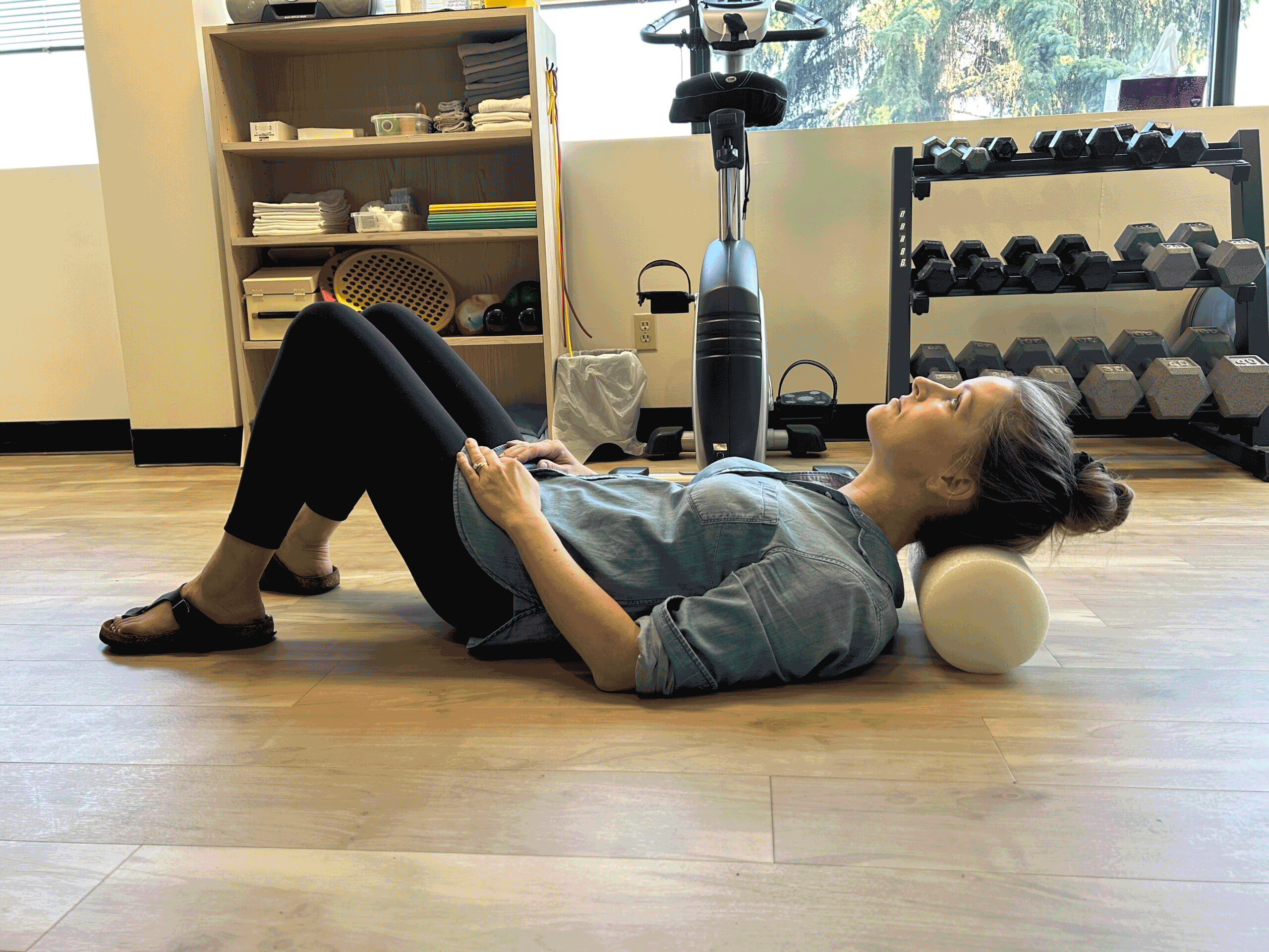 A girl laying on the floor with a foam roller behind her neck. She uses the roller to stretch out her neck up and down and on the sides.