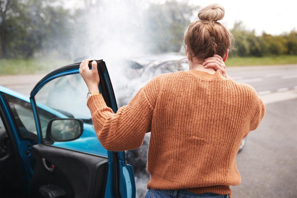 Rear view of a woman in an orange sweater standing outside a blue car after an accident, holding her neck in pain while looking at the damaged vehicle behind her. Smoke rises from the collision, indicating the severity of the impact.