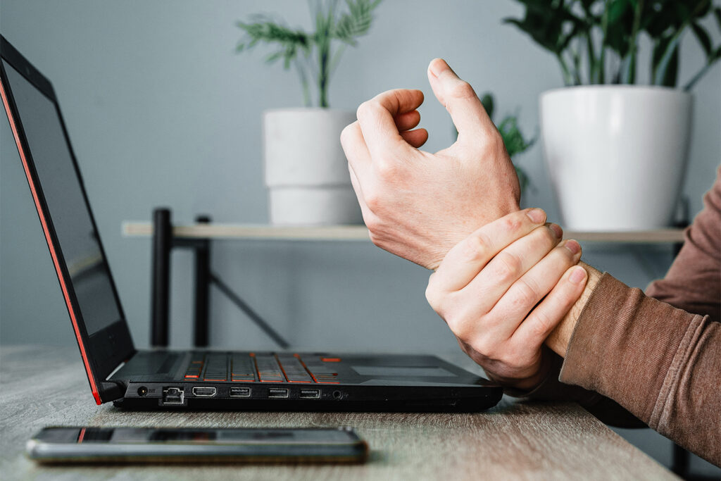 A person sitting at a desk with a laptop holds their wrist in discomfort, suggesting hand or wrist pain from computer use.